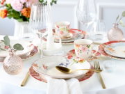 Romantisch gedeckter Tisch mit floralem Porzellan-Geschirr, goldfarbenem Besteck und geriffelten Weingläsern, fotografiert aus leicht erhöhter Perspektive schräg von vorn.