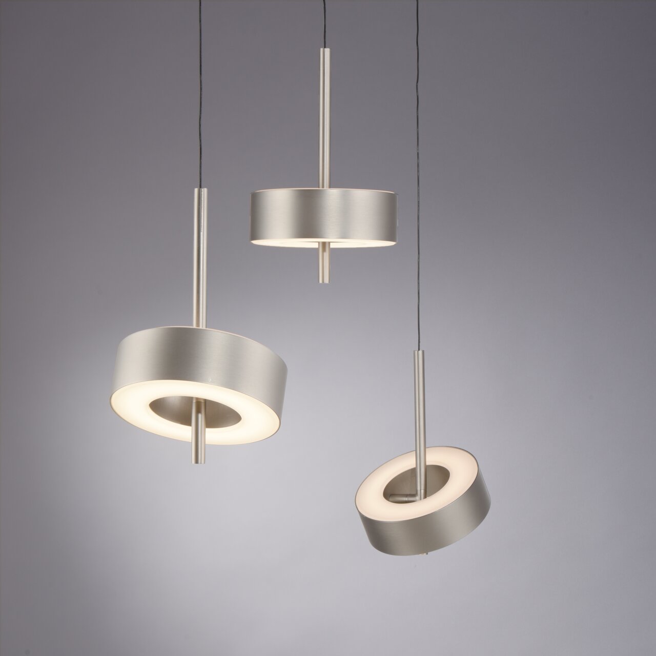 Drei moderne Pendelleuchten Q-ROTATE in gebürstetem Metall, aus seitlicher Perspektive fotografiert, mit kreisförmigem Design und warmem Licht.