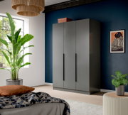 Moderner dreitüriger Kleiderschrank in Grau mit langen schwarzen Griffen, in einem Schlafzimmer vor dunkelblauer Wand, perspektivisch in leichter Frontansicht von links fotografiert
