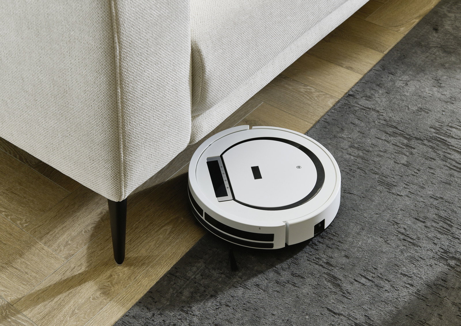 Weißer Saugroboter reinigt den Boden neben einer hellen Eckgarnitur, Perspektive von oben.