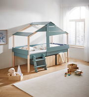 Kinderbett in Hausform mit blauer Leiter, seitliche Perspektive, umgeben von Spielzeug und einem Fenster im Hintergrund.