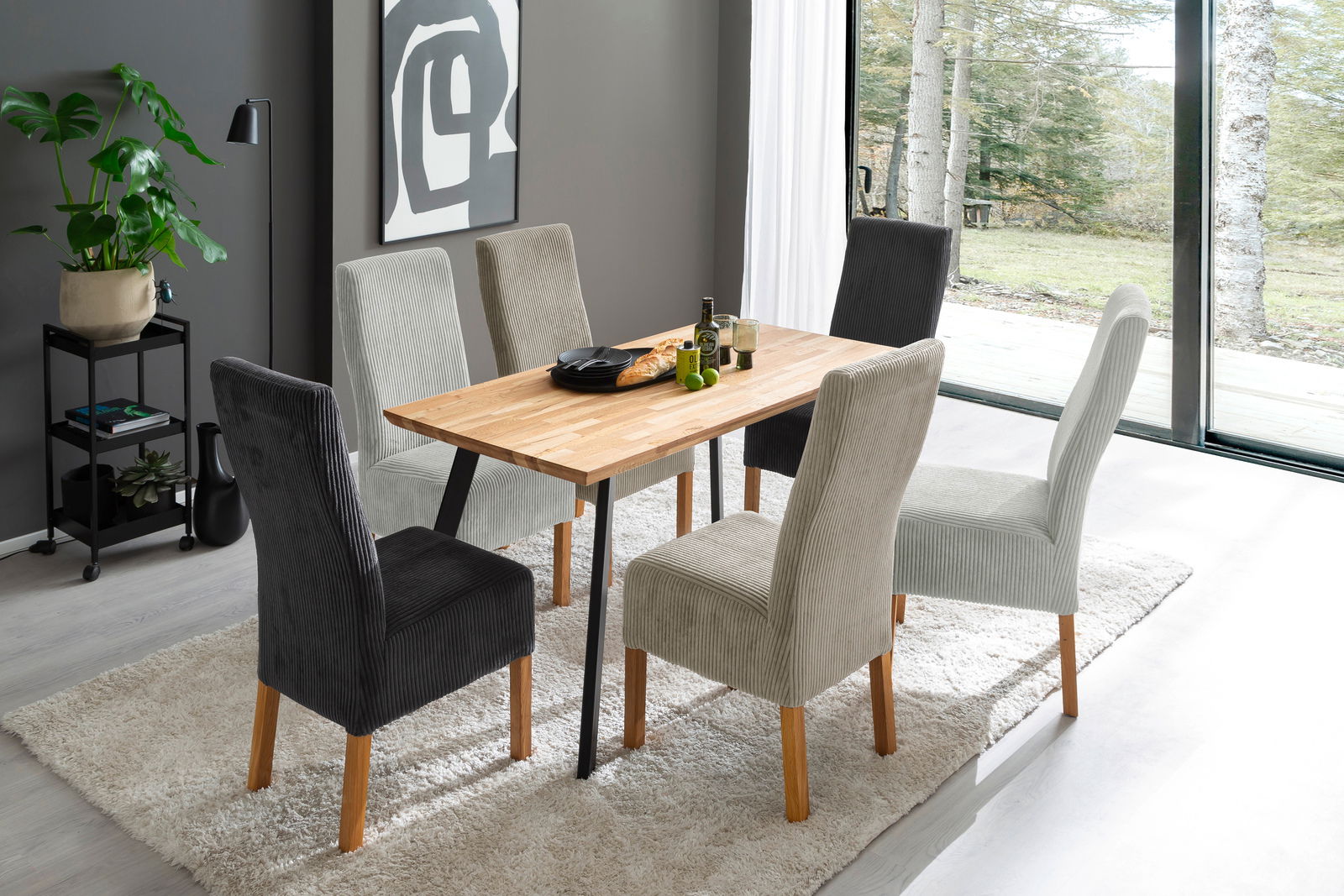 Moderner Esstisch mit Holzplatte und schwarzen Beinen, umgeben von sechs gepolsterten Stühlen in einem hellen Raum mit Blick auf den Garten. Perspektive von schräg oben.