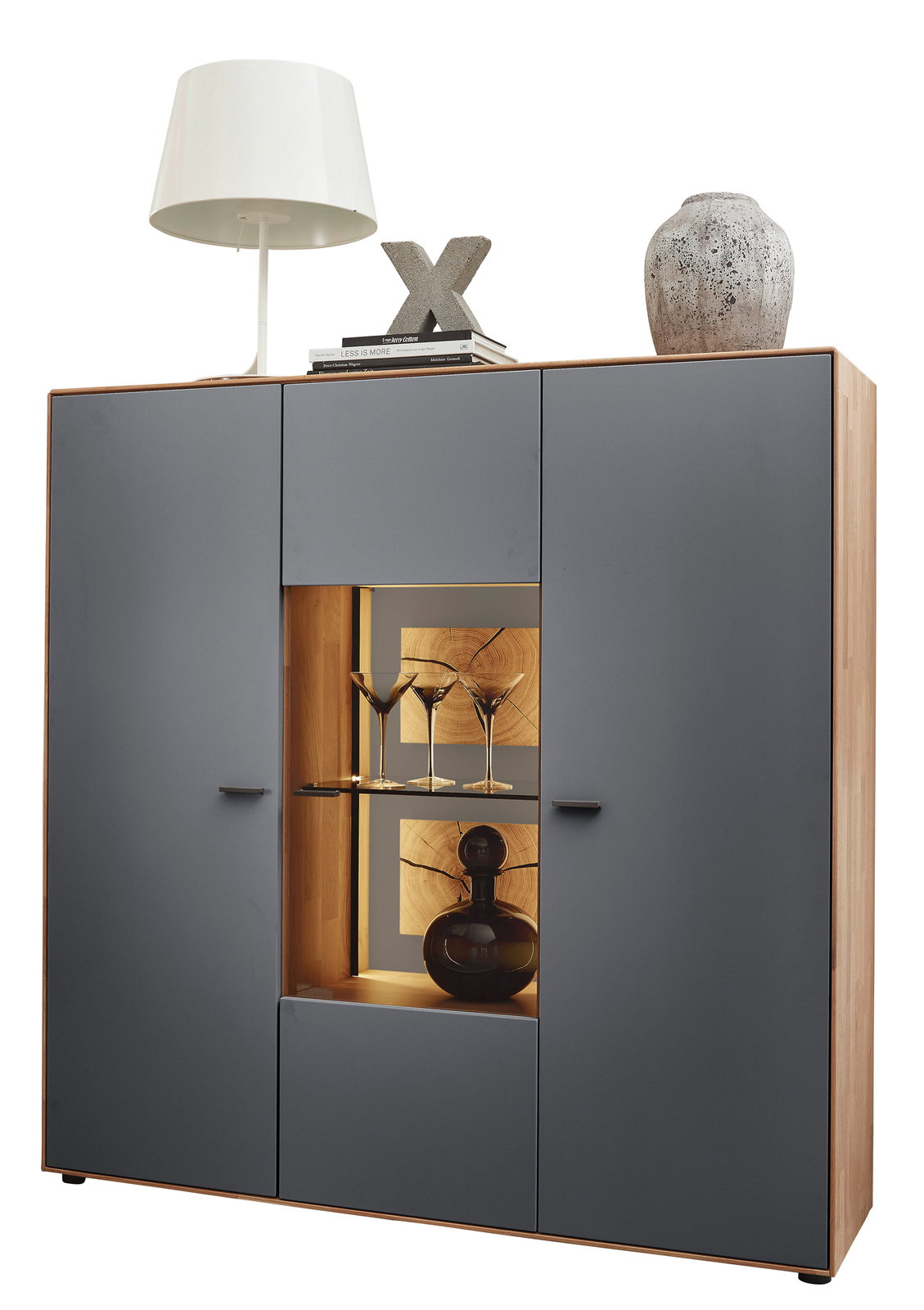 Frontale Ansicht eines modernen Highboards von Wöstmann Markenmöbel, Modell WM 2410, mit grauen Türen und einem beleuchteten Glaseinsatz in der Mitte, dekoriert mit Gläsern und einer Karaffe.