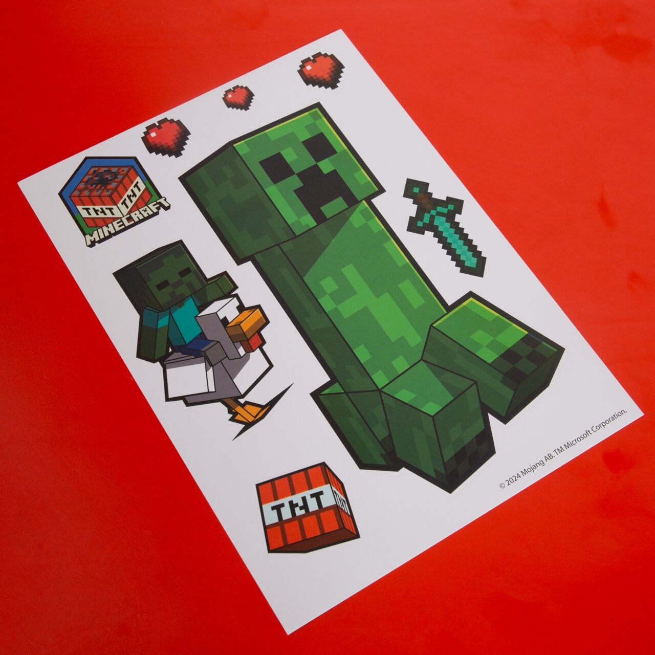 Minecraft Stickerbogen mit Creeper, Zombie auf Huhn, TNT-Block, Pixel-Schwert und Herzen auf rotem Hintergrund, schräg von oben fotografiert
