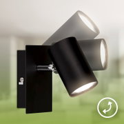 Close-up in driekwartaanzicht van een moderne zwarte wandlamp met drie verstelbare, cilindervormige spots tegen een onscherpe groene achtergrond