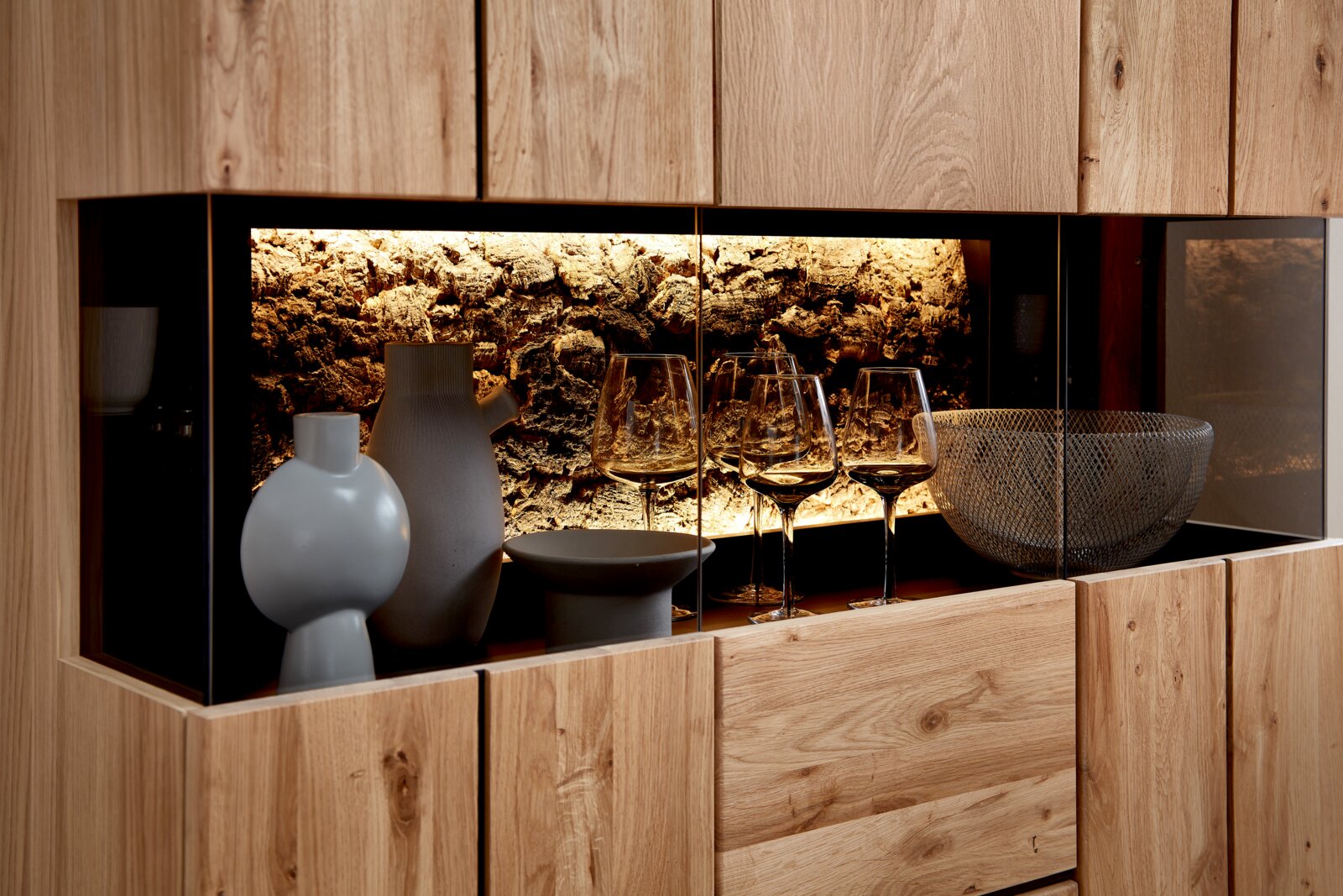 Moderne Holzvitrine mit Glasfront und warm beleuchtetem Steinrückwand-Design, dekoriert mit Keramikvasen, Schale und Weingläsern, Frontansicht leicht seitlich