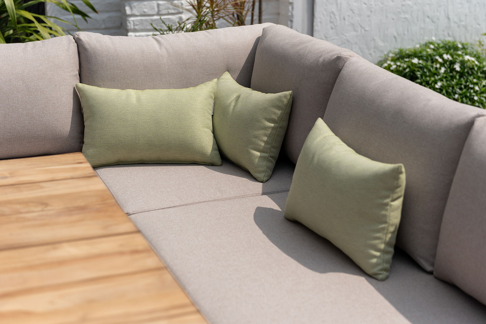 Eck-Sitzbank mit beigen Polstern und grünen Kissen, im Freien neben einem Holztisch, seitliche Perspektive