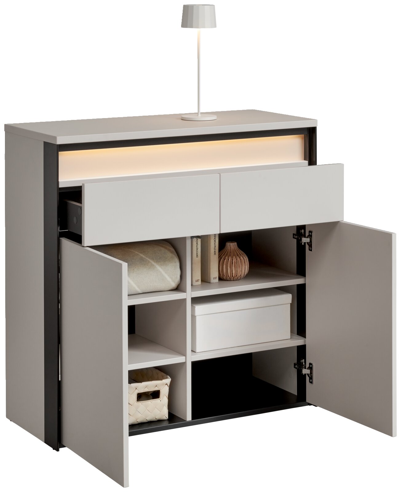 Moderne Kommode mit zwei offenen Schubladen und zwei geöffneten Türen, die verschiedene Gegenstände wie Bücher, eine Decke und eine Box zeigen. Eine Lampe steht auf der Oberseite. Perspektive: leicht von links vorne.