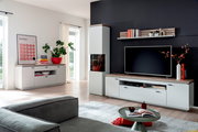 Modernes Wohnzimmer mit weiß-grauer Wohnwand: TV-Lowboard mit Holzoptik-Oberplatte, Vitrine und Sideboard sowie Wandregal vor dunkler Akzentwand, aufgenommen in schräger Frontansicht aus dem Raum.