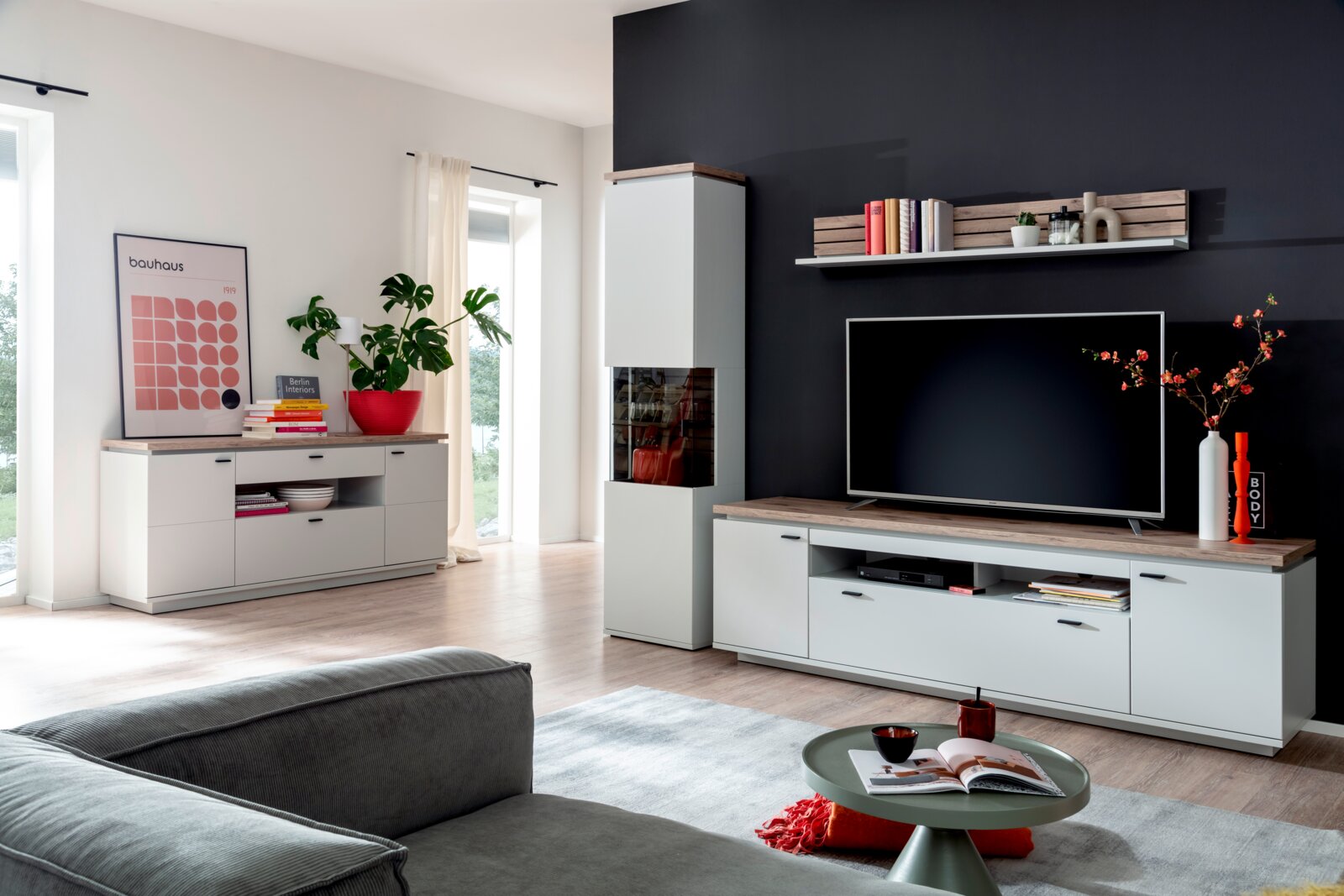 Stylife Wandcombinatie ESTEPONA Moderne woonkamer met een wit-grijze wandmeubelcombinatie: tv-lowboard met een blad in houtlook, vitrinekast en dressoir, evenals een wandrek voor een donkere accentmuur, gefotografeerd vanuit een schuin vooraanzicht vanuit de kamer.