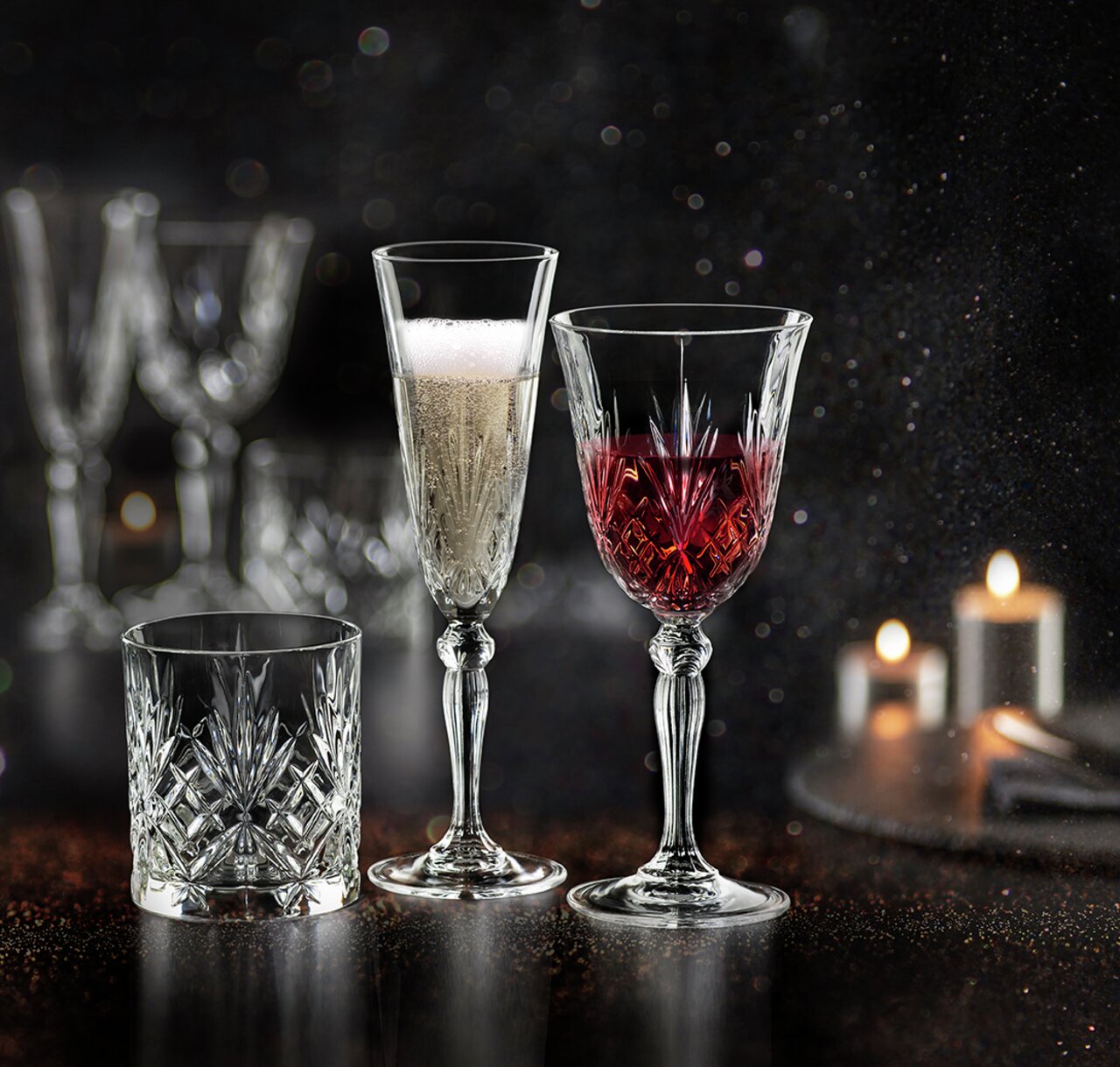 RCR Italia Servies 18-delig MELODIA Drei elegante Kristallgläser, eines mit Sekt, eines mit Rotwein und eines leer, auf einem dunklen, stimmungsvollen Hintergrund mit Kerzenlicht.