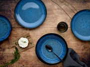 Vogelperspektive auf ein 12-teiliges Tafelservice in Blau mit rustikalem Design, bestehend aus Tellern und einer Tasse, arrangiert auf einem Holztisch.