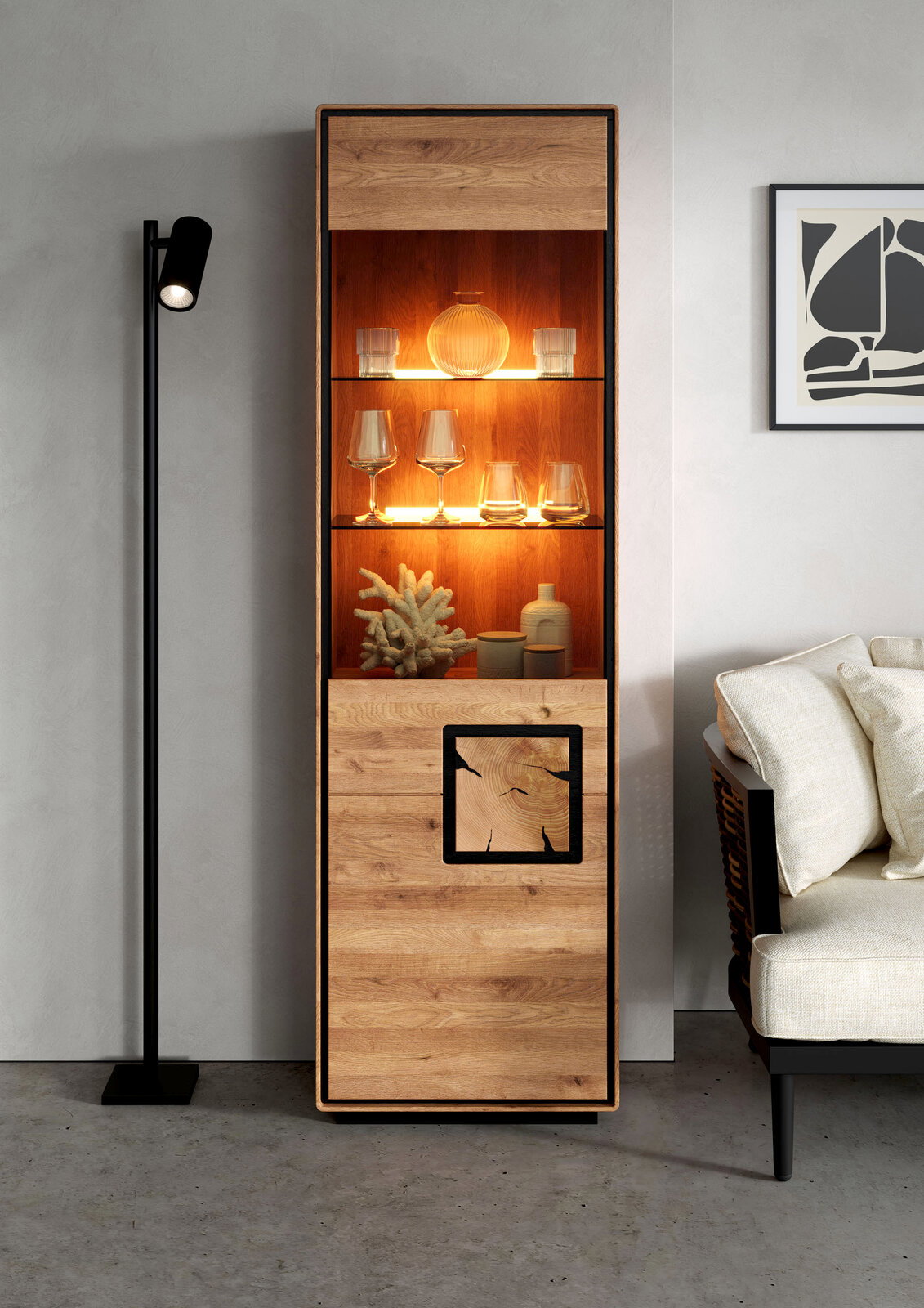 Frontale Ansicht einer hohen, schmalen Vitrine aus hellem Holz mit schwarzem Rahmen, warm beleuchteten Glasböden und dekorativem Einsatz im unteren Fach, im Wohnzimmer neben Stehlampe und Sofa