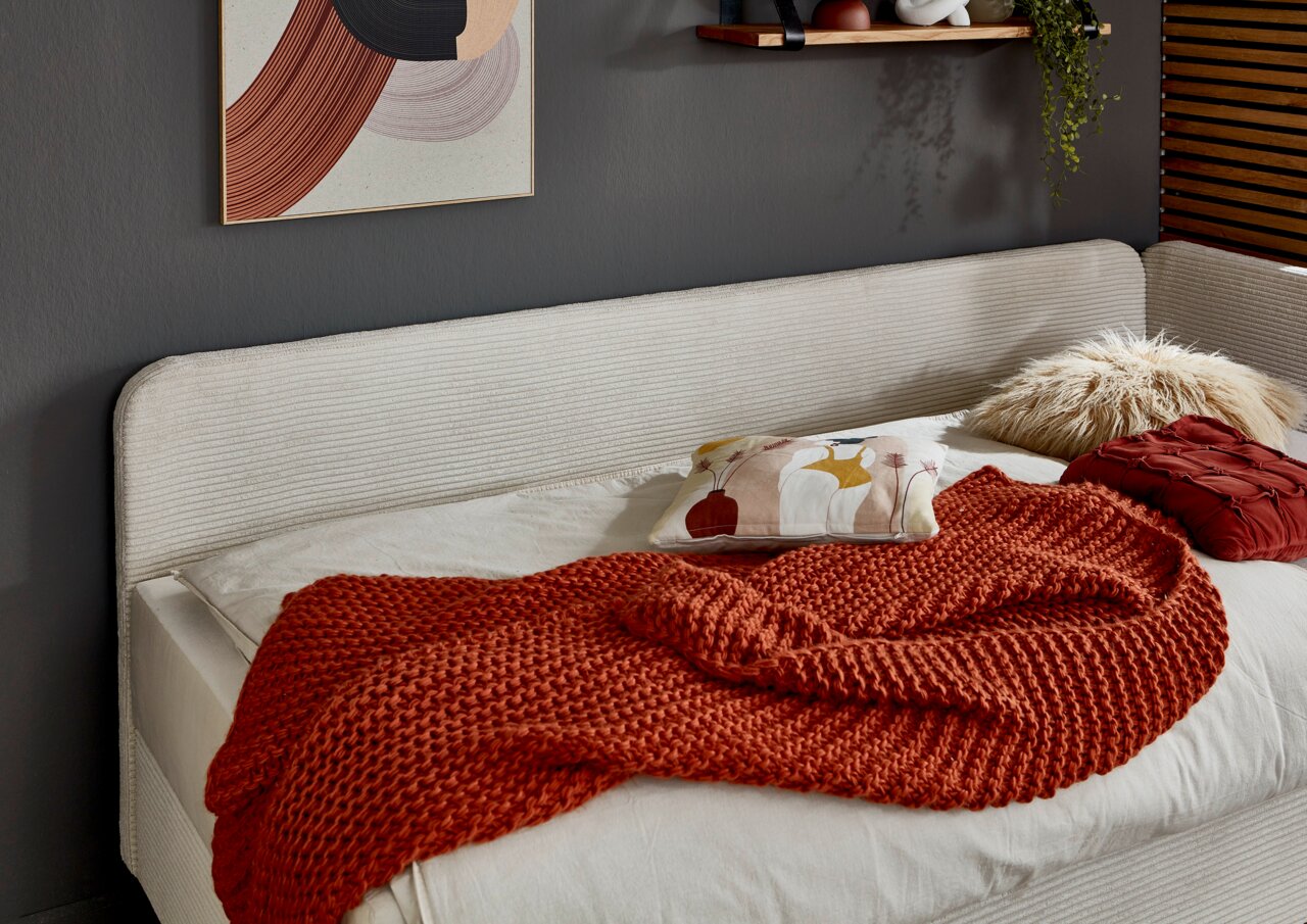 Seitliche Perspektive auf ein elegantes Polsterbett mit cremefarbenem Kopfteil, dekoriert mit einem roten Strickplaid und bunten Kissen.