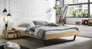 Modern laag houten bed met een zwart metalen frame, opgemaakt met grijs beddengoed en kussens in een lichte slaapkamer tegen een witte bakstenen muur; perspectief: zijaanzicht vanuit een iets verhoogde positie.