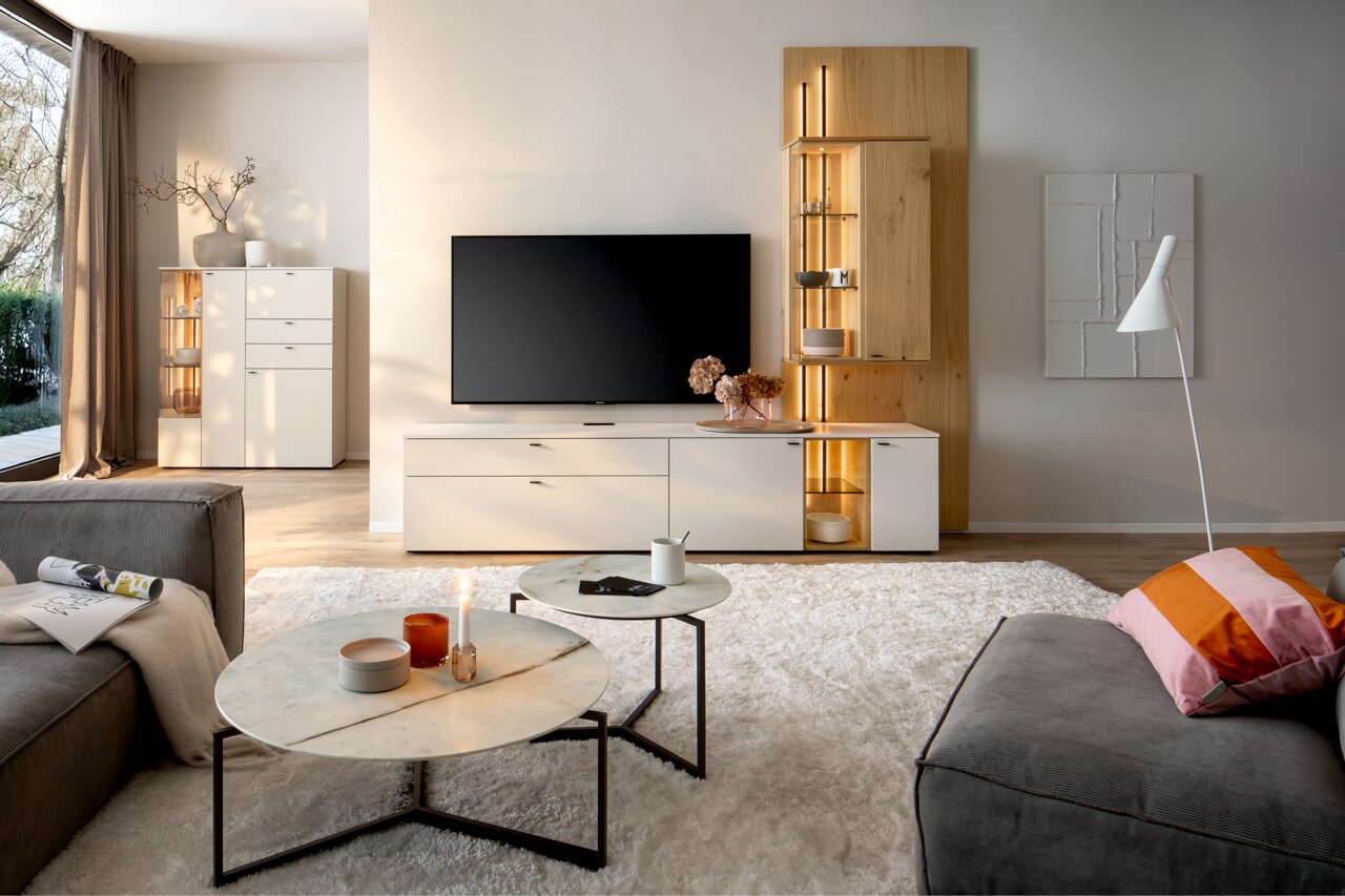 Moderne Wohnzimmer-Wandkombination mit TV, weißen Schränken und Holzakzenten, aus der Perspektive eines gemütlichen Wohnzimmers mit Sofa und Couchtischen.