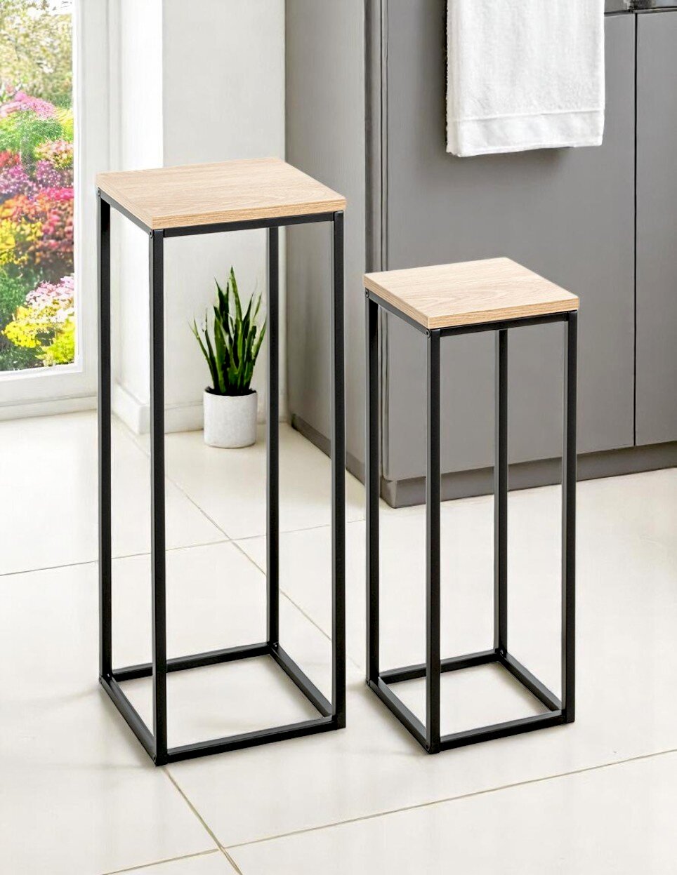 Zwei moderne Pflanzenständer/Beistelltische in unterschiedlicher Höhe mit schwarzem Metallgestell und heller Holzplatte, im Wohnraum auf Fliesenboden, Perspektive: leicht schräg von vorn (3/4-Ansicht).