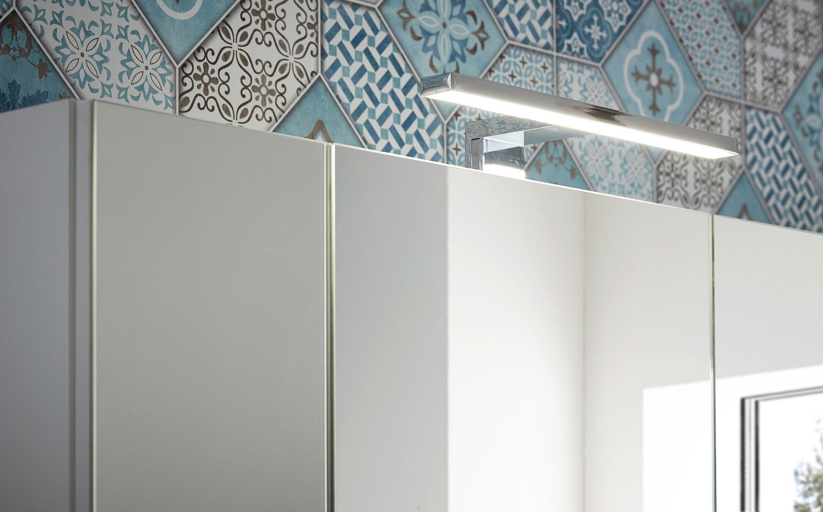 Spiegelschrank mit moderner LED-Beleuchtung, seitliche Perspektive, vor einer Wand mit blau-weißen Fliesen.