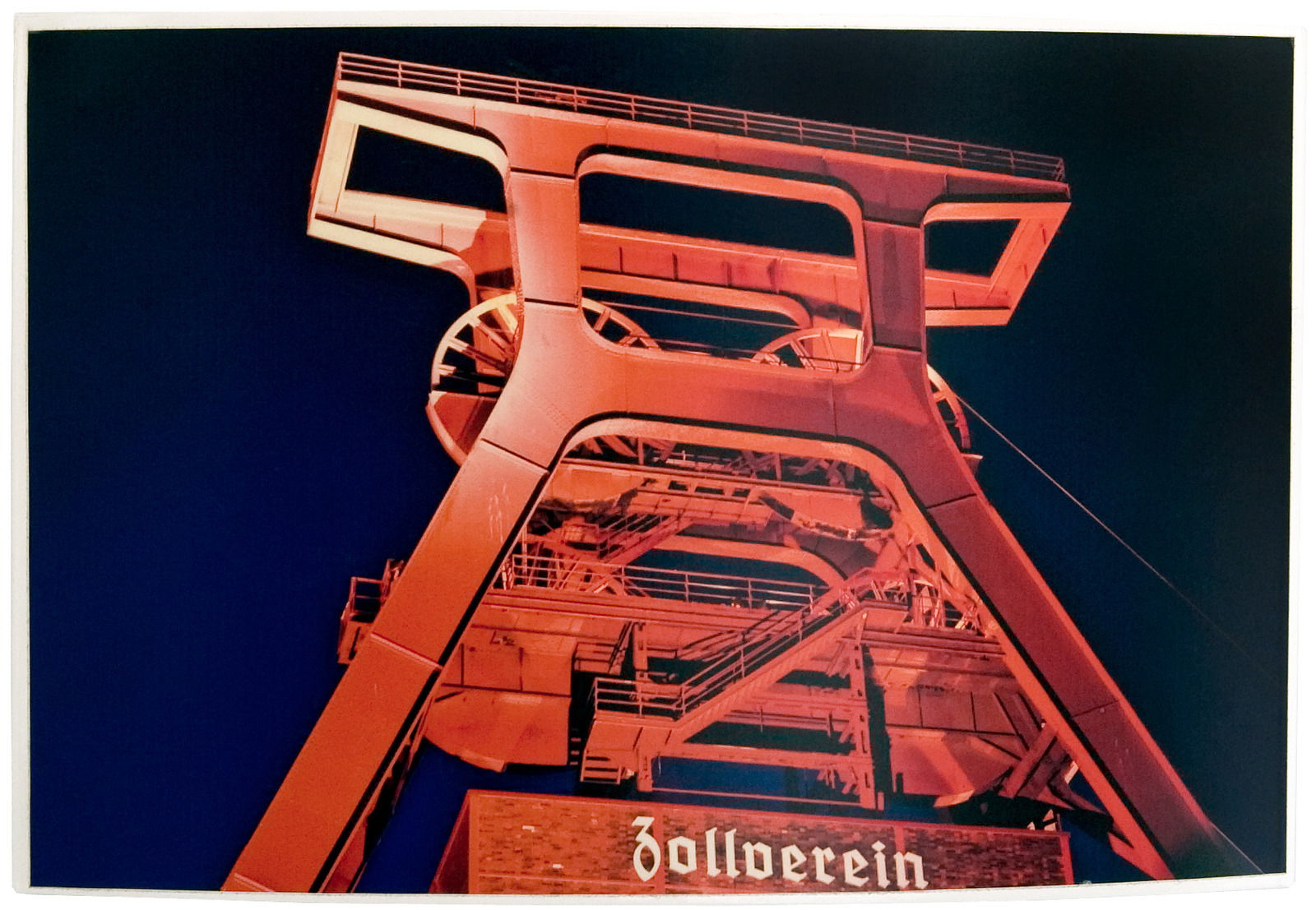 Nachtaufnahme des rot beleuchteten Förderturms der Zeche Zollverein aus einer leicht aufwärts gerichteten Perspektive.