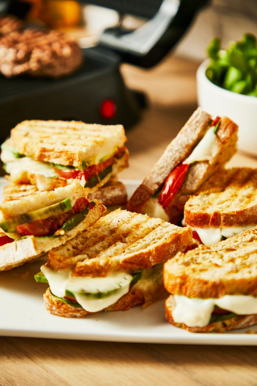 Nahaufnahme von gegrillten Sandwiches mit geschmolzenem Käse und Gemüse, im Hintergrund ein Kontaktgrill.