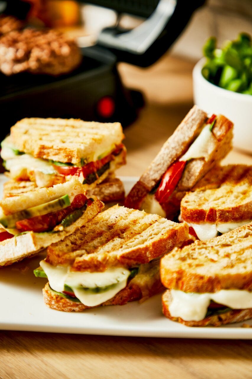 Nahaufnahme von gegrillten Sandwiches mit geschmolzenem Käse und Gemüse, im Hintergrund ein Kontaktgrill.