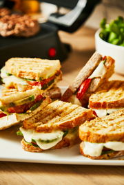 Nahaufnahme von gegrillten Sandwiches mit geschmolzenem Käse und Gemüse, im Hintergrund ein Kontaktgrill.