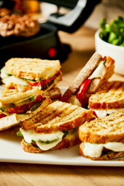 Close-up van gegrilde sandwiches met gesmolten kaas en groenten, met op de achtergrond een contactgrill.