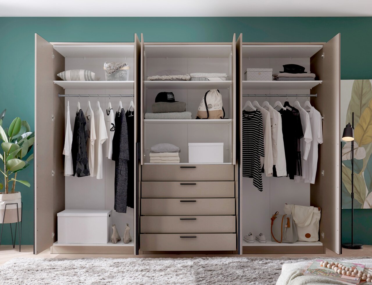 Open, moderne kledingkast in beige met drie vakken, kledingstangen, planken en vijf lades, gevuld met kleding en opbergdozen; vooraanzicht.