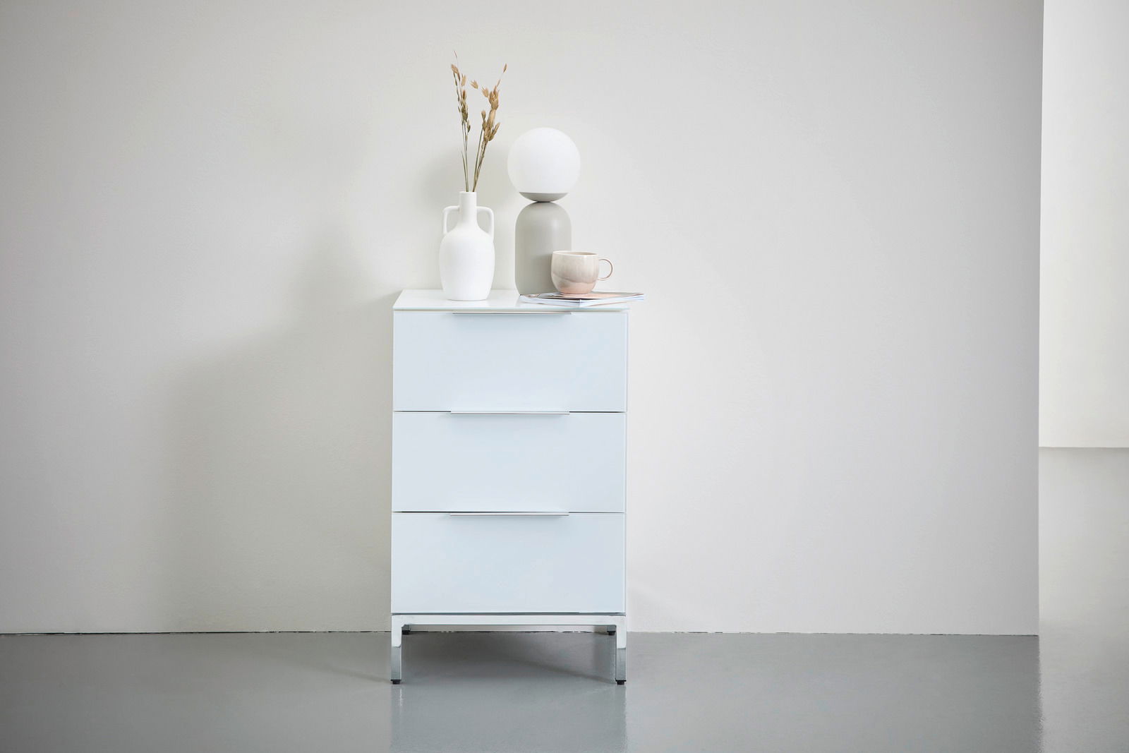 Moderne Nachtkonsole MALIN mit drei Schubladen, frontal abgebildet, dekoriert mit Vasen und einer Tasse.