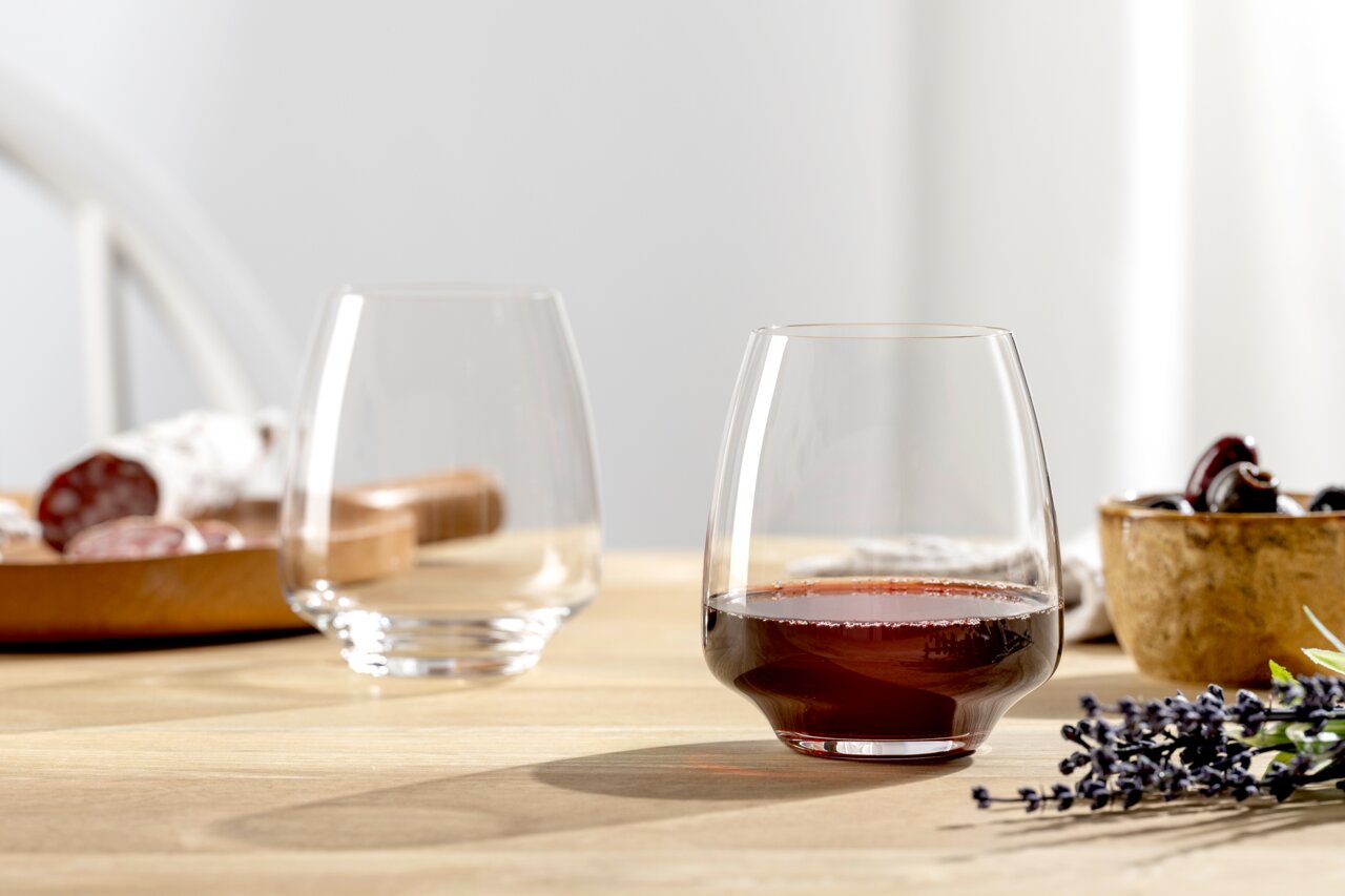 Leonardo Drinkglas CESTI Zwei elegante, bauchige Gläser auf einem Holztisch, eines gefüllt mit Rotwein, im Hintergrund eine Holzplatte mit Wurst und ein Schälchen mit Oliven, seitliche Perspektive.