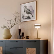 Schlichte Tischleuchte in Beige/Gold auf einem Sideboard neben Vasen und Kerzen, darüber ein gerahmtes Vogelbild an der Wand; Perspektive: leicht seitliche Ansicht von vorne