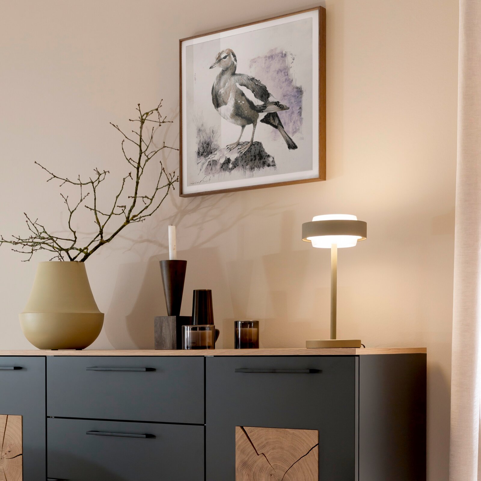 Schlichte Tischleuchte in Beige/Gold auf einem Sideboard neben Vasen und Kerzen, darüber ein gerahmtes Vogelbild an der Wand Perspektive: leicht seitliche Ansicht von vorne