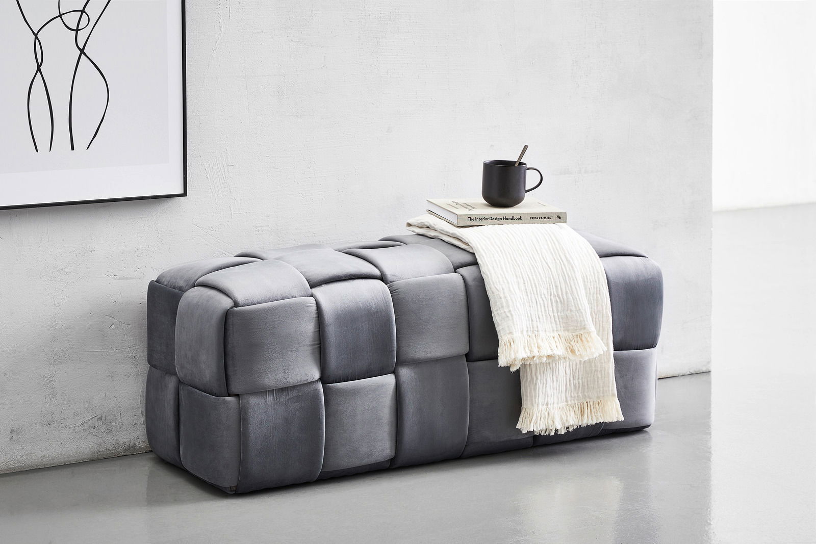 Graue Hockerbank mit gepolstertem Würfeldesign, seitliche Perspektive. Dekoriert mit einer Tasse, Büchern und einem weißen Tuch.