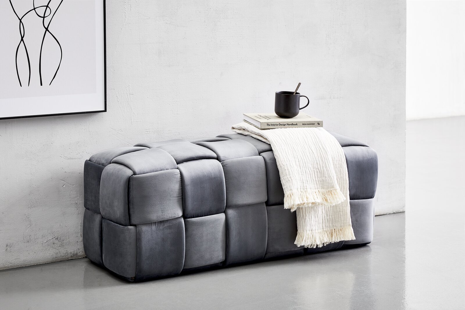 XORA Hockerbank LUCY Graue Hockerbank mit gepolstertem Würfeldesign, seitliche Perspektive. Dekoriert mit einer Tasse, Büchern und einem weißen Tuch.