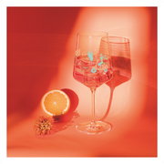 Ein Weinglas mit sommerlichem Design, gefüllt mit einem Getränk, steht auf einem orangefarbenen Hintergrund. Daneben liegt eine halbe Orange und eine Blume. Die Perspektive ist frontal.