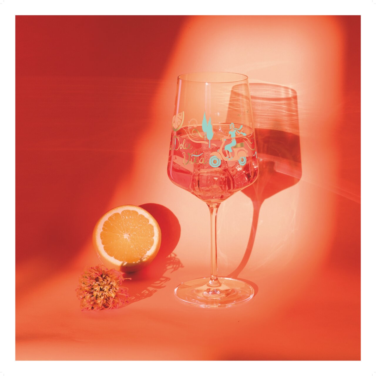 Ritzenhoff Aperizzo 4-delige set SOMMERRAUSCH Een wijnglas met een zomers ontwerp, gevuld met een drankje, staat op een oranje achtergrond. Ernaast liggen een halve sinaasappel en een bloem. Het perspectief is frontaal.