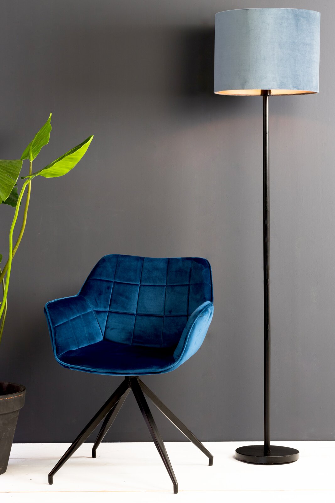 Staande lamp met een cilindervormige lampenkap in lichtblauw, op een zwarte voet, naast een blauwe fauteuil en een plant, gefotografeerd vanuit een zijdelingse hoek.