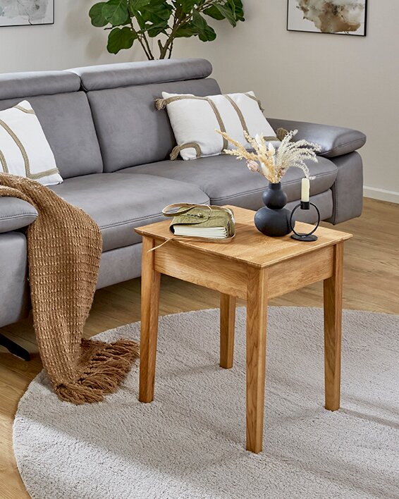 Quadratischer Beistelltisch aus hellem Holz auf rundem, hellem Teppich vor grauem Sofa, dekoriert mit Vase, Kerzenhalter und Buch, fotografiert aus leicht erhöhter, frontaler Perspektive.