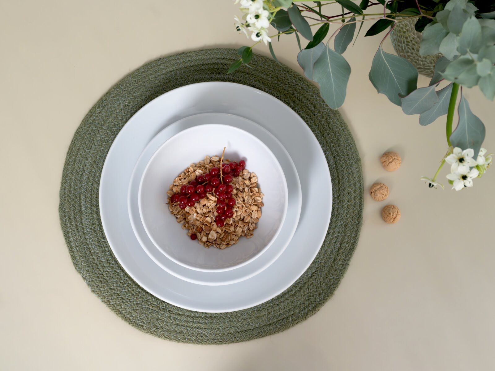Draufsicht von oben auf ein weißes Teller- und Schüssel-Set auf grünem Platzset, gefüllt mit Müsli und roten Johannisbeeren, daneben kleine Kekse und dekorative Zweige auf hellem Hintergrund