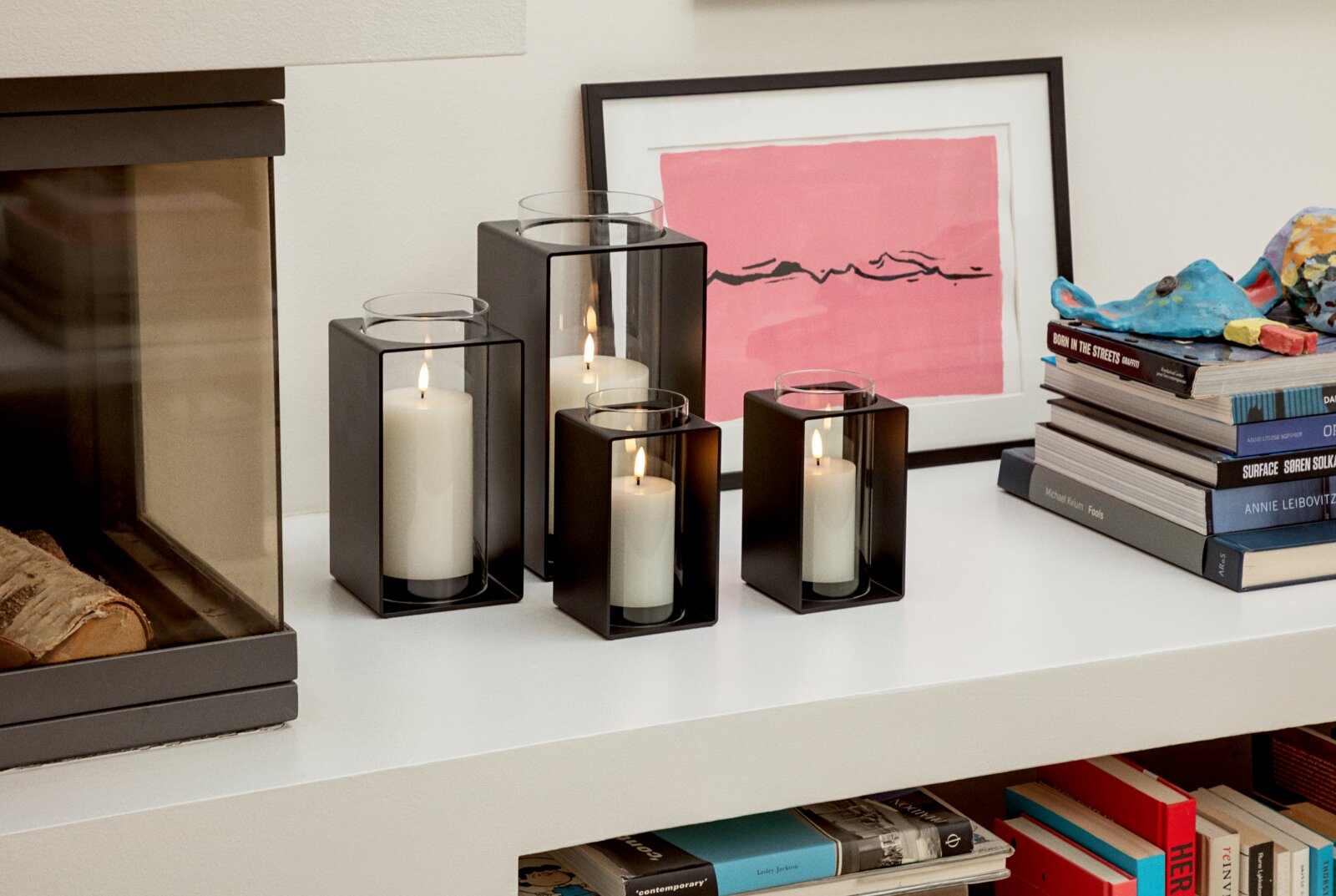 Eva Solo Glas-Windlicht 30 cm GLOW Vier moderne Windlichter aus schwarzem Metall mit Glaszylinder und brennenden weißen Stumpenkerzen in unterschiedlichen Größen auf einem weißen Regal neben einem gerahmten Bild und einem Bücherstapel, Perspektive: frontale leicht seitliche Ansicht.