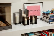 Vier moderne Windlichter aus schwarzem Metall mit Glaszylinder und brennenden weißen Stumpenkerzen in unterschiedlichen Größen auf einem weißen Regal neben einem gerahmten Bild und einem Bücherstapel, Perspektive: frontale leicht seitliche Ansicht.