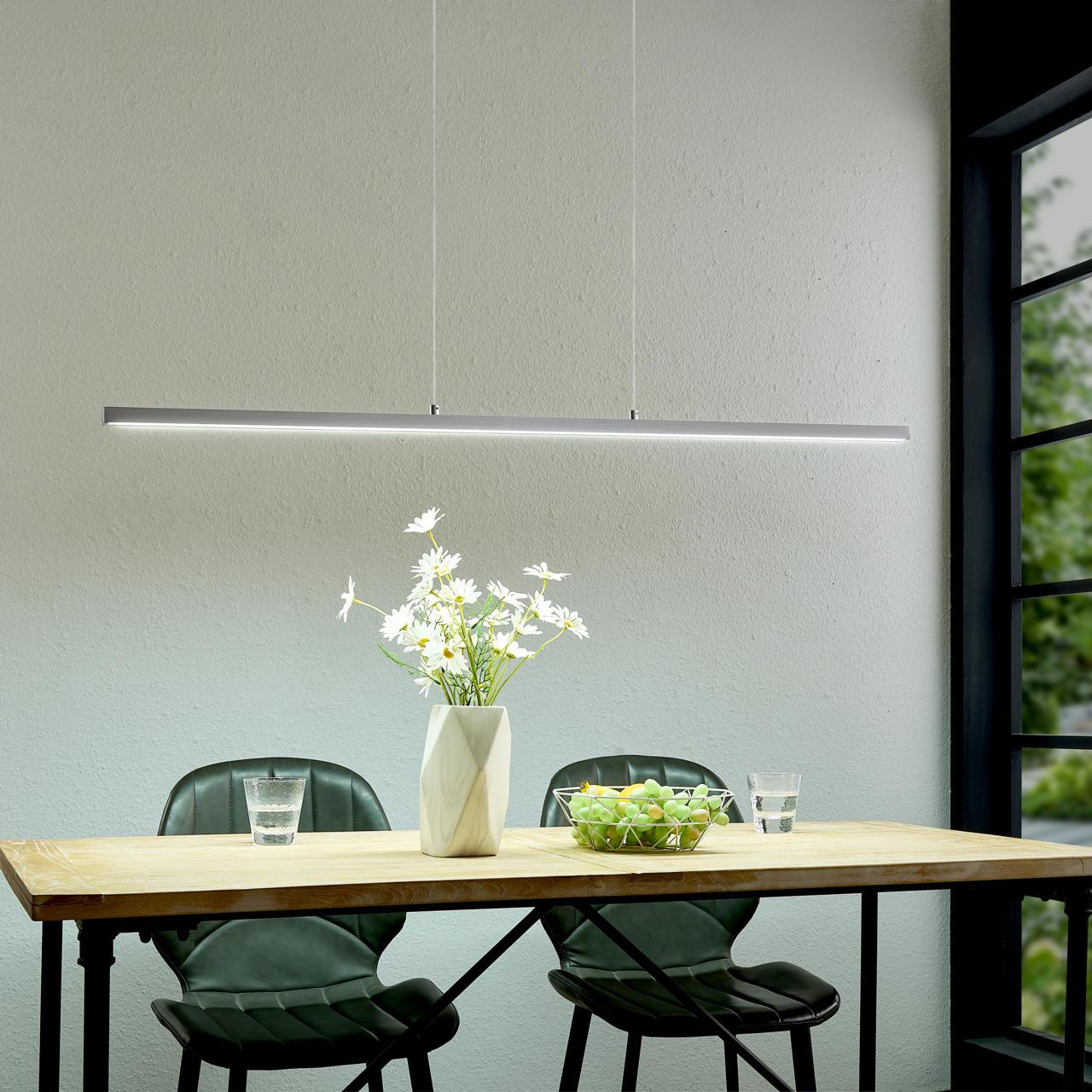 Moderne, schlanke LED-Pendelleuchte in Silber über einem Holztisch mit zwei grünen Stühlen, Vase mit weißen Blumen und Obstschale; Perspektive: frontale Ansicht auf Augenhöhe.
