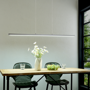 Moderne, schlanke LED-Pendelleuchte in Silber über einem Holztisch mit zwei grünen Stühlen, Vase mit weißen Blumen und Obstschale; Perspektive: frontale Ansicht auf Augenhöhe.