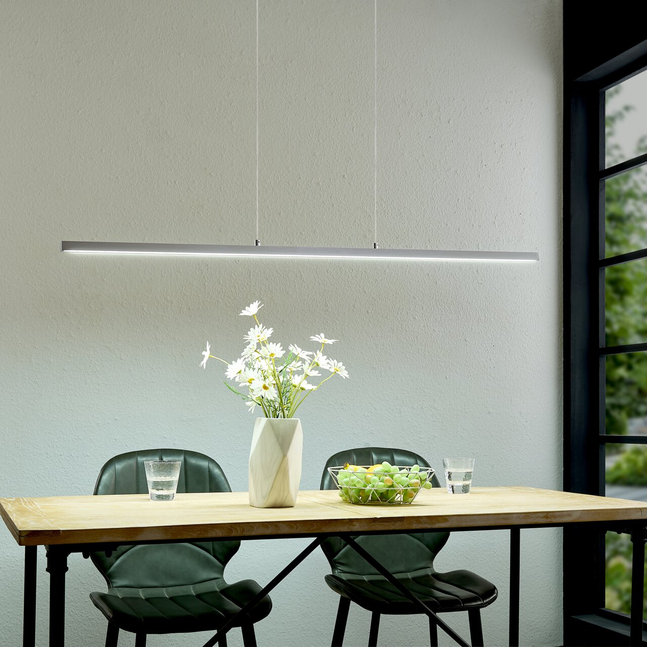 Moderne, slanke LED-hanglamp in zilver boven een houten tafel met twee groene stoelen, een vaas met witte bloemen en een fruitschaal; perspectief: vooraanzicht op ooghoogte.
