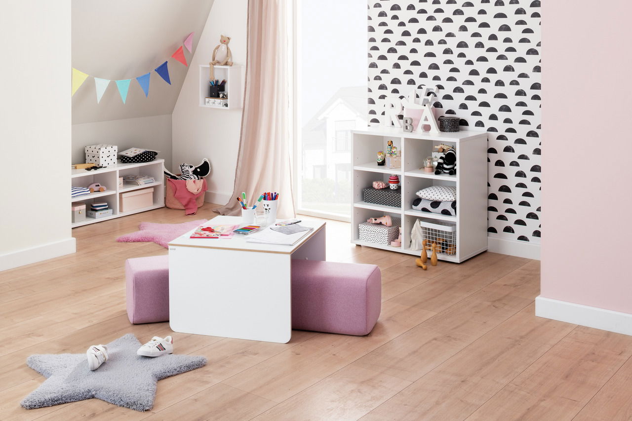 Kinderzimmer mit einem weißen Kindertisch und einem rosa Sitzpolster, umgeben von Regalen mit Spielzeug und Dekorationen. Perspektive von vorne.