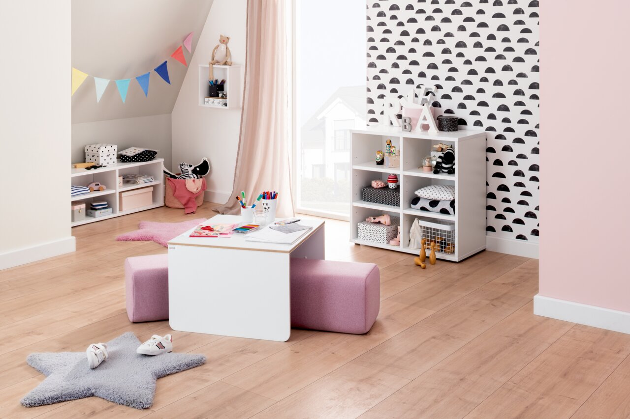 Kinderzimmer mit einem weißen Kindertisch und einem rosa Sitzpolster, umgeben von Regalen mit Spielzeug und Dekorationen. Perspektive von vorne.
