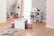 Kinderzimmer mit einem weißen Kindertisch und einem rosa Sitzpolster, umgeben von Regalen mit Spielzeug und Dekorationen. Perspektive von vorne.