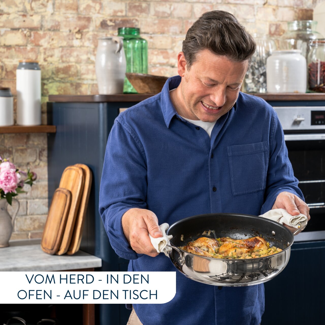 Mann in blauer Jacke hält eine Servierpfanne mit gebratenem Gericht in einer Küche, seitliche Perspektive.