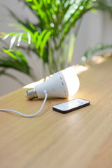 Akku-Leuchtmittel auf einem Tisch liegend, mit angeschlossenem Ladekabel und Fernbedienung, seitliche Perspektive