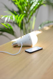 Akku-Leuchtmittel auf einem Tisch liegend, mit angeschlossenem Ladekabel und Fernbedienung, seitliche Perspektive